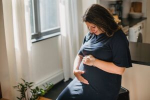 Positive pregnant female with brown hair in dress sitting on chair and touching belly in cozy apartment in daytime